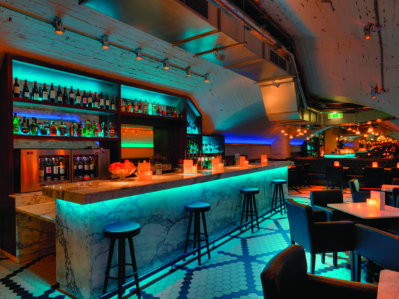 An image of the Alston's white, marble bar. The floor is tiled in small black and white tiles, laid in geometric shapes. The bar and the white, brick walls are up-lit with turquoise lights. 4 minimal black stools sit along the length of the bar, while small, low marble tables are surrounded by modern, brown leather armchairs to the right of it. Shelves line the curving wall behind the bar, they are also lit turquoise and are well stocked with bottles and glasses.