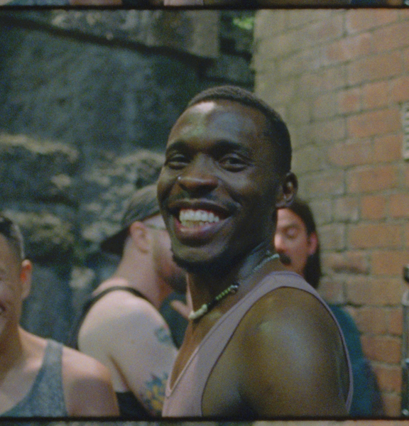 A young man in a vest widely smiling looking directly at the camera, with other young men smiling in the background