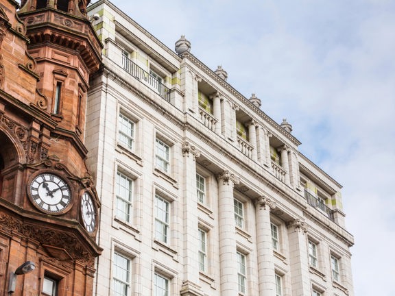 An exterior view of the Native Apartments Glasgow building depicts the top 4 storeys of a grand, white Victorian building, against a cloudy sky. The image has been taken from below. Urns, pillars and balustrades adorn the facade and top of the building. Large, white, sash and case windows can be seen on each floor. The corner of another Victorian building, this time in red sandstone, can also be seen in the foreground. It has 2 clock faces and is adorned with carved finials, gables and an angular turret.