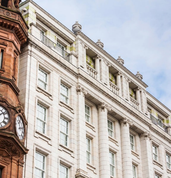 An exterior view of the Native Apartments Glasgow building depicts the top 4 storeys of a grand, white Victorian building, against a cloudy sky. The image has been taken from below. Urns, pillars and balustrades adorn the facade and top of the building. Large, white, sash and case windows can be seen on each floor. The corner of another Victorian building, this time in red sandstone, can also be seen in the foreground. It has 2 clock faces and is adorned with carved finials, gables and an angular turret.