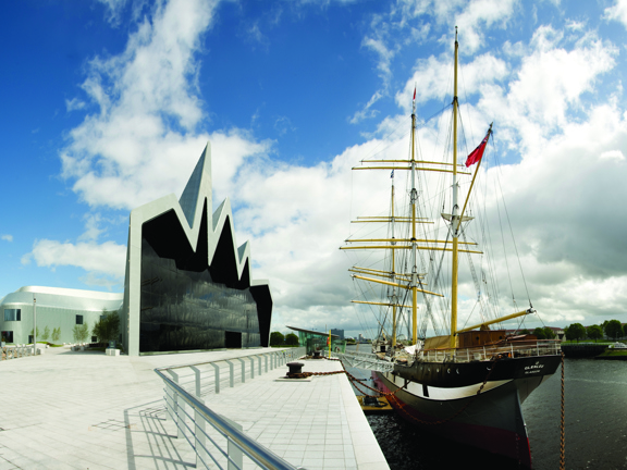 An exterior of the Riverside Museum. On the left, the museum is a modern silver building with a cartoonish, zig-zagging roofline. The main facade if a huge, dark glass wall which overlooks the Tall Ship and the River Clyde. Outside the building is a wide, pale stone, paved area and footpath with a silver railing between the building and the waters edge. A ramps leads to the Tall Ship is moored on the right, its hull is black and white and its multiple masts are bright yellow against the blue cloudy sky.