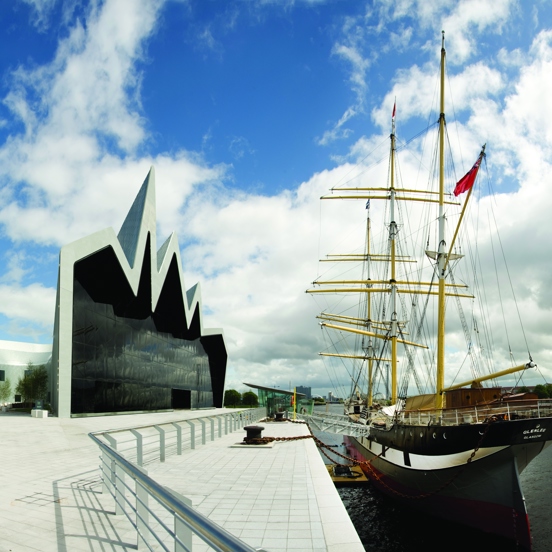 An exterior of the Riverside Museum. On the left, the museum is a modern silver building with a cartoonish, zig-zagging roofline. The main facade if a huge, dark glass wall which overlooks the Tall Ship and the River Clyde. Outside the building is a wide, pale stone, paved area and footpath with a silver railing between the building and the waters edge. A ramps leads to the Tall Ship is moored on the right, its hull is black and white and its multiple masts are bright yellow against the blue cloudy sky.