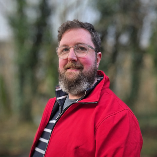 A smiling man with glasses and a full beard, wearing a bright red jacket outdoors against a softly blurred woodland background.