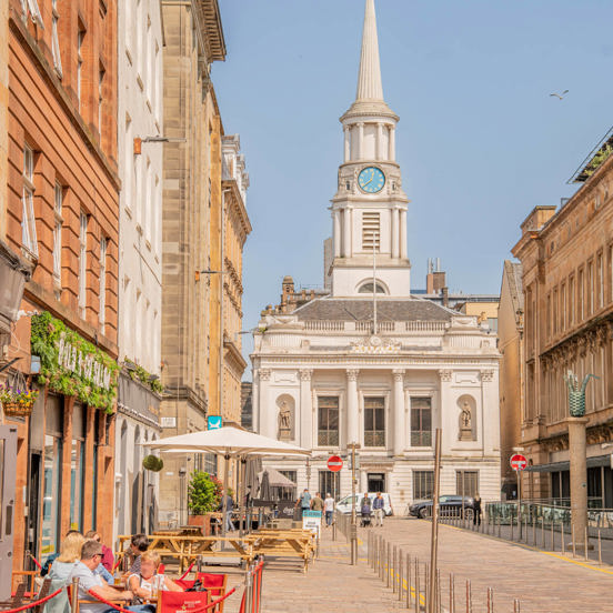 sunny city scene with people dining outdoors and spired building at the end of the street