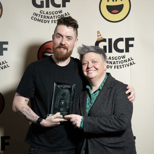 Comedian Rosco McClelland stands in front of Comedy festival backdrop hugging female comedian Susie McCabe while holding Sir Billy Connolly Spirit of Glasgow award