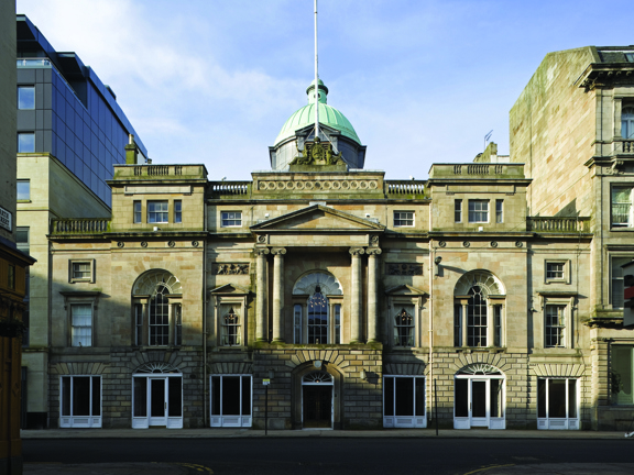 An exterior of the Glasgow Trades Hall shows a 3 storey, symmetrical, Victorian sandstone building. It has carved details, including ionic columns either side of the central, 1st floor arched window and a carved coat of arms finial on top. A large verdigris dome and a flagpole also adorn the top of the building. The image is taken from over the road, a level pavement can be seen outside of the building's entrance. Buildings flank the Trades Hall on either side.