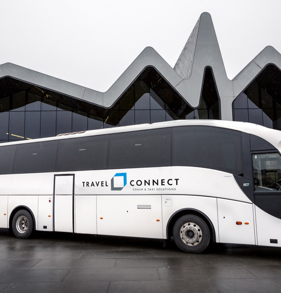 An image of a large white coach with the Travel Connect logo on the side. The coach is parked on the concourse outside of the Glasgow Riverside Museum, with its distinctive zig-zagging roofline. The floor is paved and wet from rain.