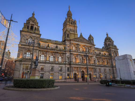 Front view of the outside of Glasgow City Chambers. 2 statues on plinths surrounded by hedges can be seen in the foreground, in front of the building.