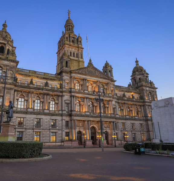 Front view of the outside of Glasgow City Chambers. 2 statues on plinths surrounded by hedges can be seen in the foreground, in front of the building.