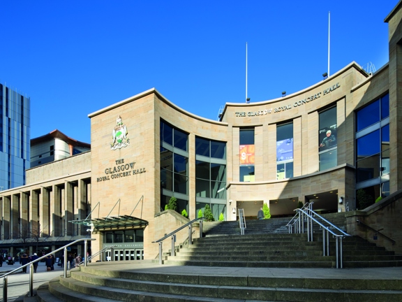 An exterior image of the venue. Curved steps with metal handrails lead up to the curved stone facade of the concert halls main entrance. The side of the building bears the Glasgow coat of arms and lettering reading, "The Glasgow Royal Concert Hall". The venue has 3 large doorways at the top of the stairs and floor to ceiling windows flank them over 3 storeys. There are 2 flag poles on top of the building seen against a clear, blue sky. Modern high-rise buildings can be seen to the left of the image.