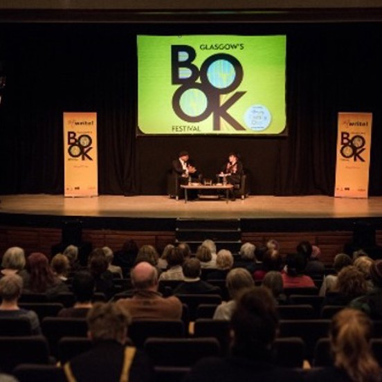 An audience sit in a theatre, on stage two people seated talking in front of a sign that reads Glasgow's Book Festival.