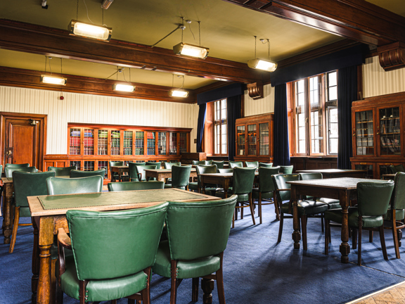A traditional room with wooden beams overhead and built in wooden bookshelves with glass doors. There are several wooden tables each with 4 green leather chairs around them sat on a blue carpet. 