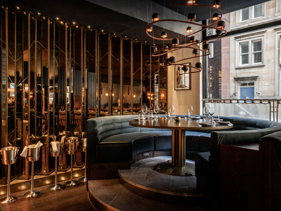 An image of a circular dining table set for 6, surrounded by a large circular booth, there are 2 carpeted steps up into the booth, the floor of the main restaurant is wood effect. The booth's bench is upholstered in grey/ blue velvet and the table is brassy. The booth is positioned in a corner, where the plate glass window meets a polished, golden, mirrored feature wall. A modern, twisting light feature is mounted to the ceiling, centrally above the table.