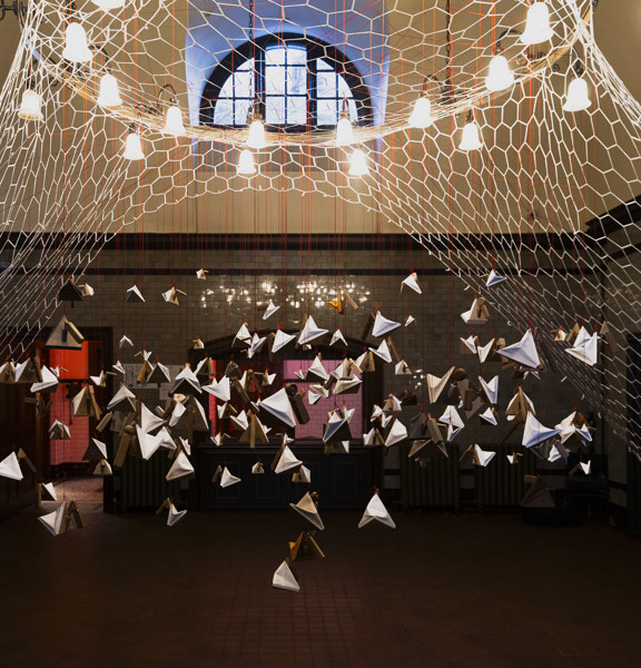 Objects in a triangular shape hang from a circular net in the middle of the ceiling, with light bulbs above them