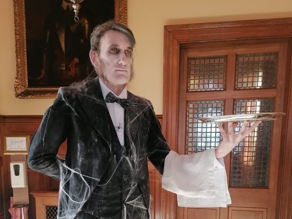 Person dressed as a male butler wearing a black suit with white shirt and bowtie. The man is draped in cobwebs and holds a silver tray in one outstretched arm with a white napkin across the arm.  