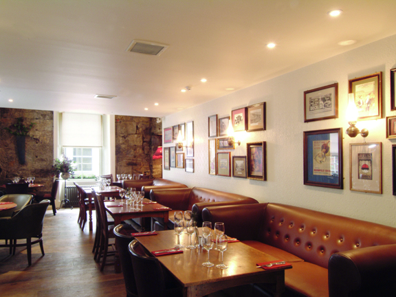 An interior photo of The Bothy restaurant shows a room with wooden floors. The room is mainly lit by daylight streaming through a large, sash and case window with white frames in the far wall. The far wall is bare sandstone while the others are plastered and painted white, with groups of framed prints and brass sconces adorning them. Plush, brown leather benches line the right-hand wall with wooden dining tables and chairs beside them. The tables have silverware and wine glasses arranged on them.